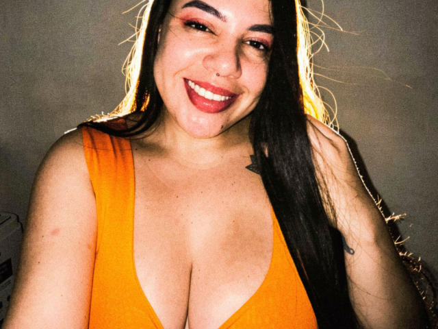 Andrea Busty, com longos cabelos pretos, sorrindo, vestindo um vestido laranja com decote profundo, mostrando seios grandes, batom vermelho, piercing no nariz e uma pequena tatuagem de estrela no lado direito do peito..