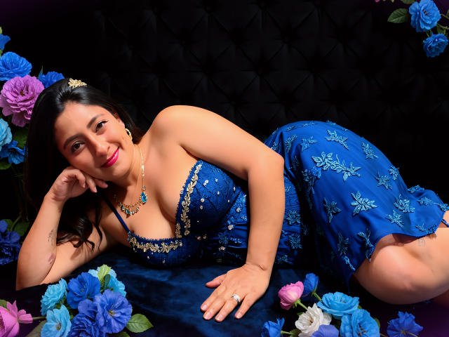 MoraMoon lying on blue velvet, wearing a blue dress with gold embroidery, showing her tits, surrounded by blue and pink roses, smiling with pink lipstick, black hair.
