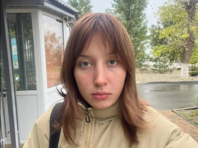 RosaleeDickey with brown hair, green eyes, and a neutral expression, wearing a beige jacket, standing near a glass booth, carrying a black backpack.