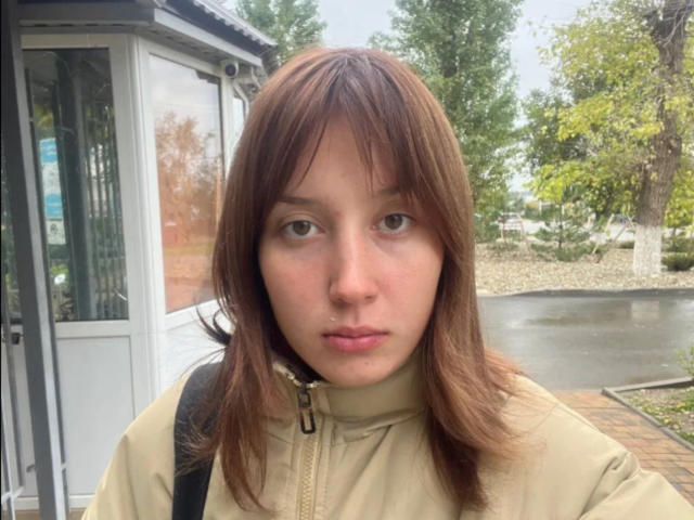 Rosalee Dickey tiene el cabello castaño, ojos verdes y una expresión neutra. Lleva una chaqueta beige con cremallera y está de pie frente a un escaparate de vidrio..