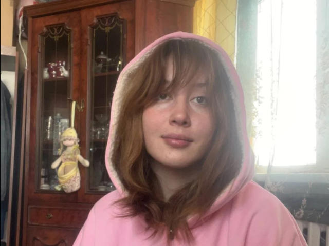 Rosalee Dickey, com moletom rosa, cabelo castanho ondulado e um sorriso discreto, usando o capuz, com uma boneca em um armário de vidro atrás dela. Suas nádegas não estão visíveis..