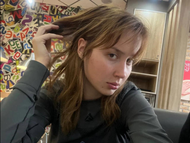 RosaleeDickey with brown hair, looking sideways, holding hair with one hand, wearing a gray long-sleeve shirt, pouty lips, slightly dirty skin.