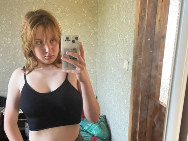 RosaleeDickey with medium-sized tits in a black sports bra, holding a phone, taking a mirror selfie, looking slightly annoyed, with light brown hair, in a textured room.