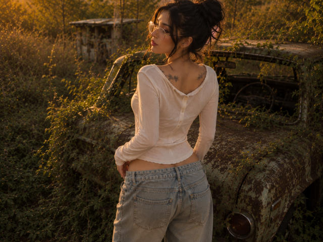 Celeste Ocampo, dos au dos de la caméra, portant une chemise blanche à manches longues et un jean bleu, se tient devant une vieille voiture couverte de mousse, montrant ses fesses et ses tatouages..
