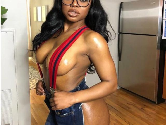 LiaSquirt with shiny brown skin, large bare boobs, and dark nipples, wearing red suspenders, blue jeans, and glasses, standing in a kitchen with a stainless steel fridge.