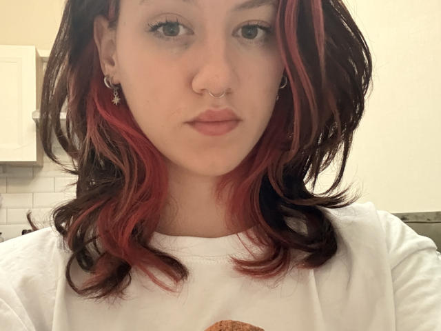JeannineNorals ha i capelli rossi e neri, un piercing al naso e orecchini a forma di stella, indossa una camicia bianca, con un'espressione neutra e un trucco leggero..