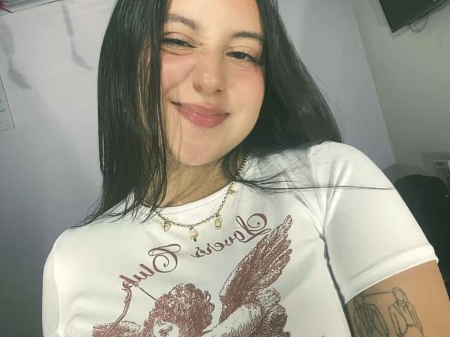 Rosalie Dupont heeft lang zwart haar, een lichte glimlach en draagt een witte blouse met een bruine engelprint. Ze heeft een gouden ketting en een tatoeage op haar arm..