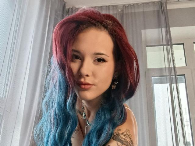 RachelNervis con capelli a sfumatura rossa e blu, seno piccolo, tatuaggio sulla spalla destra, che guarda la fotocamera, indossa orecchini a forma di stella, in piedi davanti a tende trasparenti, con luce proveniente dalla finestra..