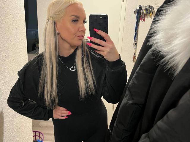 MiaXXX - attraente, con capelli biondi, vestito nero, unghie rosa, collana a forma di cuore, che si scatta un selfie allo specchio, con una mano appoggiata al fianco, giacca nera con pelliccia bianca sul lato destro..