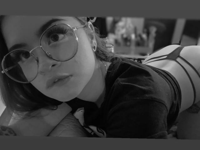 RavenThuwu con gafas, acostada en una cama, vistiendo una camiseta negra y tangas negras con tirantes, mostrando su trasero, mirando a la cámara con un ligero puchero. Foto en blanco y negro..