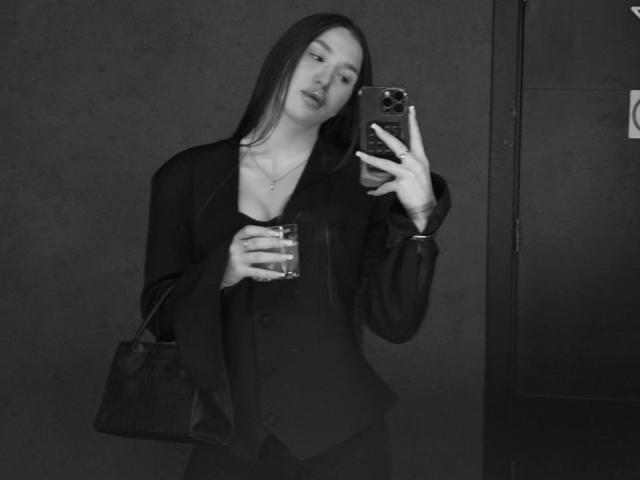Ingeborg Wheadon, con lunghi capelli scuri, tiene il telefono nella mano destra, indossa un blazer nero, una camicia abbottonata e una collana, tiene un bicchiere nella mano sinistra, e ha una borsa nera sulla destra..