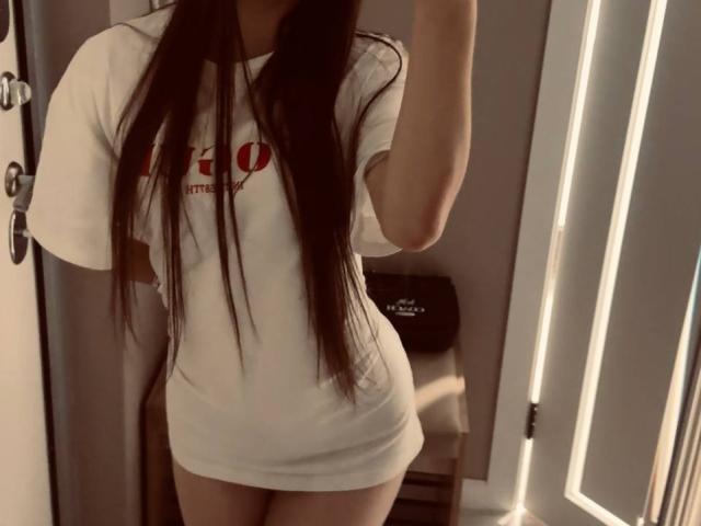 Ingeborg Wheadon, vestida con una camiseta blanca, con el pelo largo y castaño cubriendo sus glúteos, de pie en un marco de puerta, selfie tomada con un espejo, brazo derecho levantado, puerta blanca ligeramente visible a la derecha..