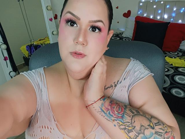 GiiannaMorrys with pink eyeshadow, nose ring, and pink lipstick, wearing a sheer white top, showing her tattoos on her right arm and hand on a chair.