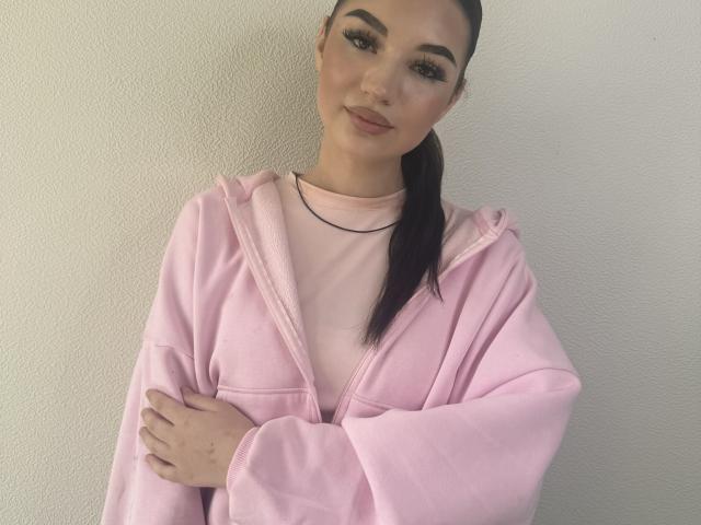 Dominique Maravilla, con una felpa rosa, le braccia incrociate e un leggero sorriso, indossa una collana nera e una maglietta rosa, e ha lunghi capelli scuri raccolti in una coda di cavallo..
