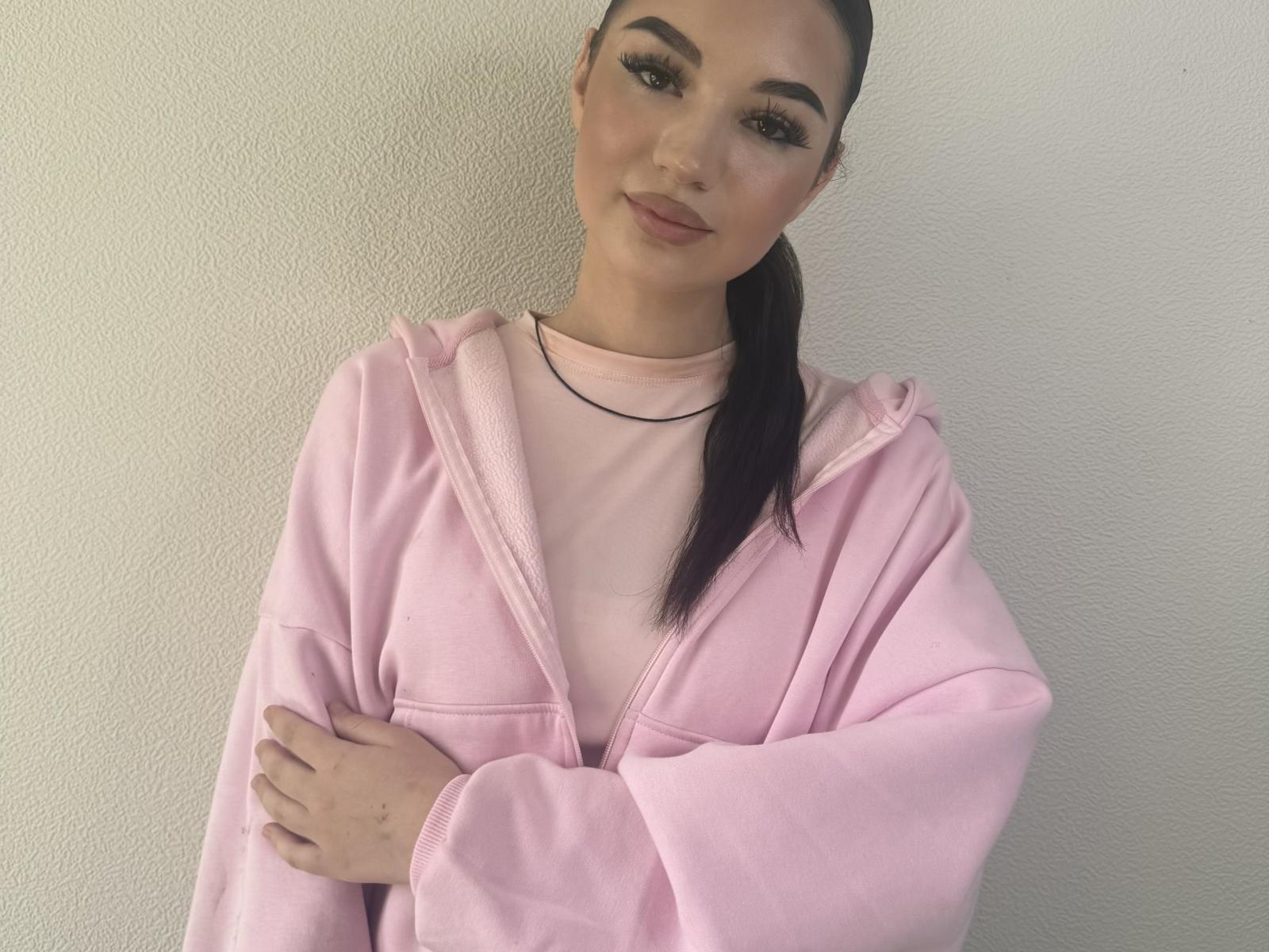Dominique Maravilla, portant un sweat à capuche rose, les bras croisés et un léger sourire, porte un collier noir et une chemise rose, et a de longs cheveux noirs attachés en queue de cheval..
