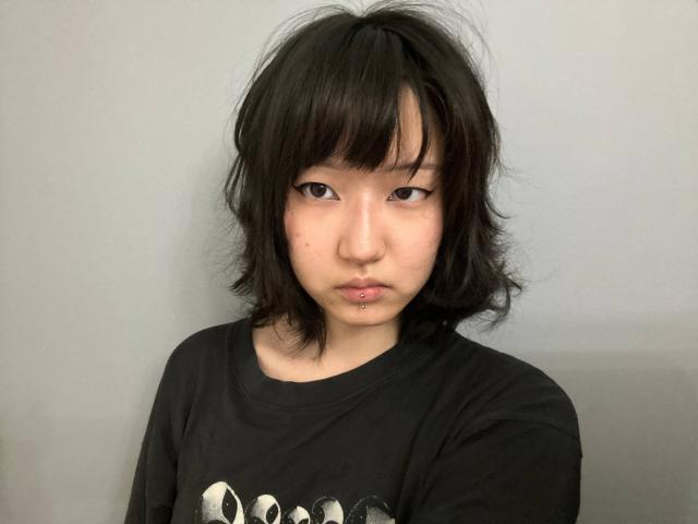 Kayce Molinas, com cabelo preto e desalinhado, pele clara e um piercing no lábio, vestindo uma camisa preta e com uma expressão ligeiramente irritada. Suas nádegas pequenas e arredondadas estão parcialmente visíveis..