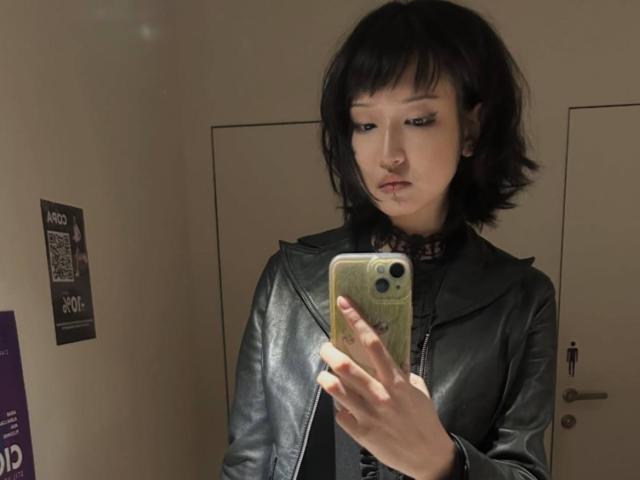 Kayce Molinas, com cabelo preto curto, vestindo uma jaqueta de couro preta, tirando uma selfie no espelho com um telefone verde, olhando para baixo com uma expressão neutra..