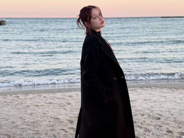 Bubochka with red hair, black coat, standing on sandy beach, looking back, hands in pockets, ocean in background, sunset sky, gentle waves, calm, confident, sexy, subtle.