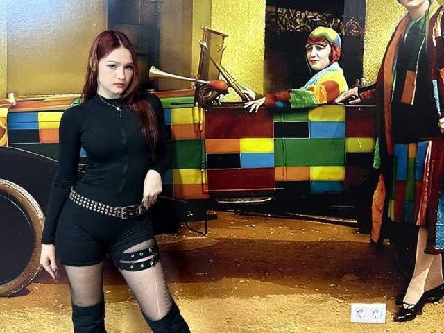 Bubochka with red hair, black zipped dress, belt with studs, and thigh strap, standing by colorful, checkered car. Another Bubochka with colorful headwear and dress in background.