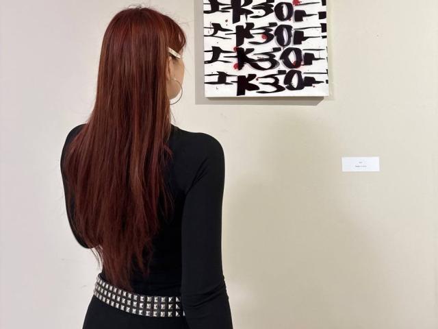 Bubochka with long red hair, black long-sleeve top, and studded belt, standing in front of abstract black-and-white painting. Bubochka's back is to the camera.