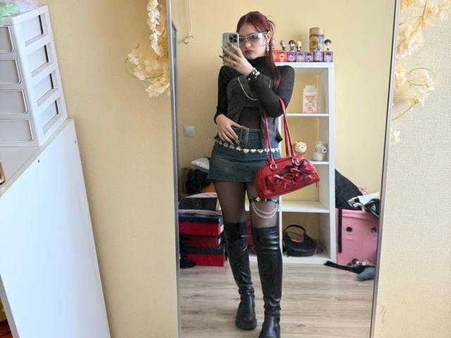 Bubochka mit roten Haaren, Brille, schwarzem Oberteil, grünem Rock, schwarzen Stiefeln, einer roten Tasche, steht vor einem Spiegel, hält ein Handy und zeigt in einem Selfie ihren Hintern und ihre Brüste..