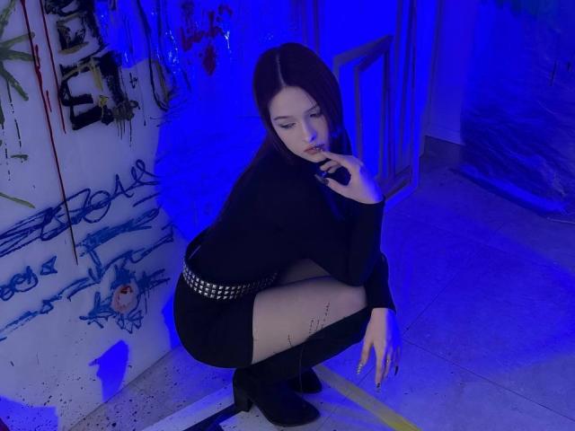 Bubochka, hockend, schwarzes Outfit, Gürtel mit silbernen Nieten, schwarze Strumpfhosen, schwarze Stiefel, blaues Licht, Finger an den Lippen, langes, dunkles Haar, blasse Haut, verführerischer Blick, Graffiti-Wand im Hintergrund..