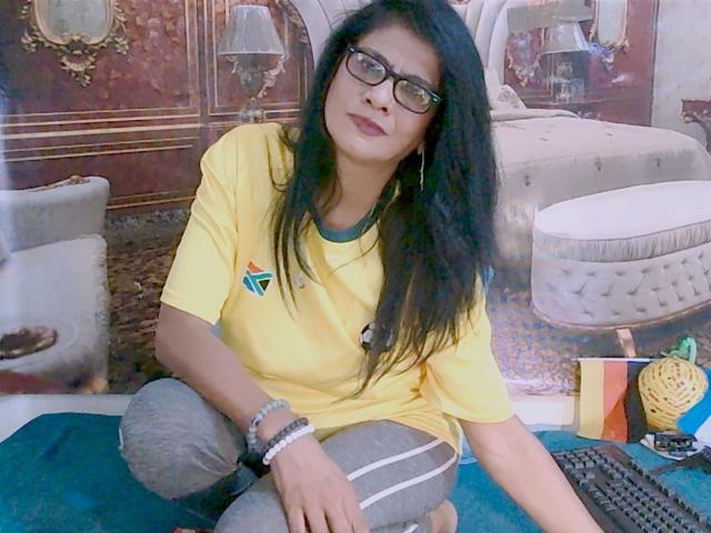 IndianAllure69 est assis en tailleur, portant une chemise jaune et un pantalon gris, avec de longs cheveux noirs et des lunettes, et il porte un bracelet, devant un clavier..