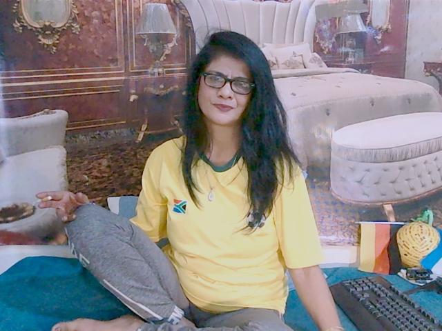 IndianAllure69 est assis en tailleur, portant une chemise jaune et un pantalon gris, avec de longs cheveux noirs et des lunettes, tenant un oreiller, et un clavier sur un tapis bleu..