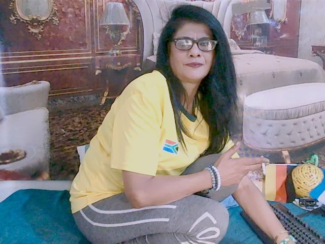 IndianAllure69, avec de longs cheveux noirs, des lunettes, une chemise jaune, un pantalon gris, assise sur un tapis bleu turquoise, tenant une cigarette, portant des bracelets, souriant légèrement..