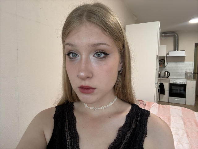 OpalMccullars hat helle Haut, langes blondes Haar, blaue Augen mit starkem schwarzem Eyeliner, rosafarbenen Lippenstift und trägt eine schwarze Spitzenbluse mit einer silbernen Kette um den Hals. Ihre Brüste sind klein..