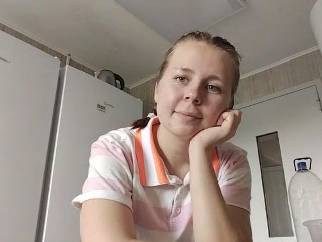 Johnetta Urtiaga, avec de larges seins, des yeux marron et un léger sourire, reposant la tête sur sa main, portant une chemise polo rose et blanche, dans une cuisine..