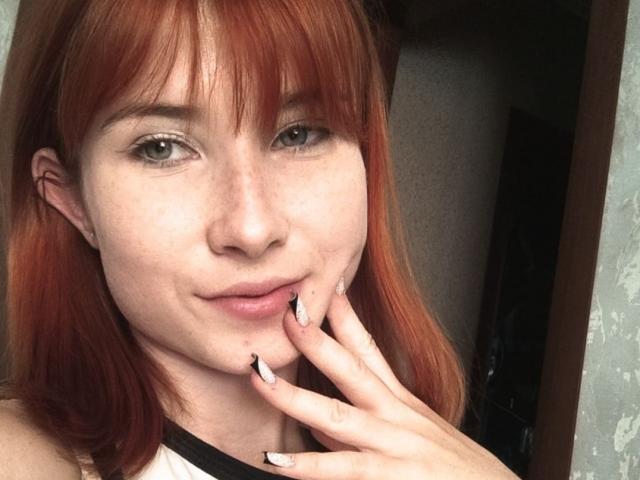 Kasandra Grothe, met rood haar, groene ogen en sproeten, die haar vinger bijt met zwarte en witte nagellak, en een lichte glimlach. Haar hand is dichtbij haar gezicht..