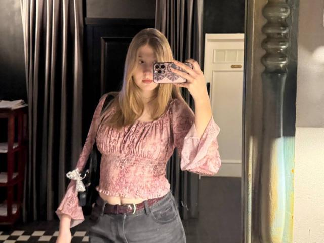 Opal McCullars mit langen blonden Haaren, einem pinkfarbenen Oberteil mit Blumenmuster und grauen, hochgeschnittenen Jeans, hält ein Telefon mit einer blau gemusterten Hülle vor einem Spiegel..