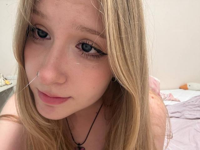 OpalMccullars tem olhos azuis, cabelos loiros longos e um nariz pequeno com um piercing de prata. Ela está usando um colar preto, e seus lábios estão ligeiramente entreabertos..