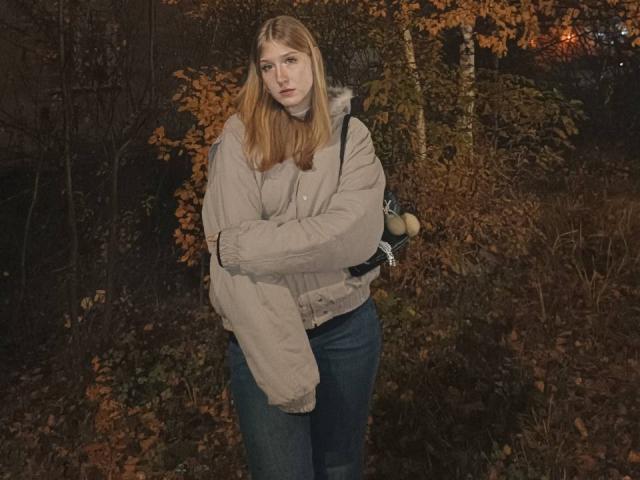 Opal McCullars, com cabelos loiros longos, vestindo uma jaqueta bege, jeans azuis e uma mochila preta, está em pé em uma área escura e arborizada, com folhas de outono. Ela parece séria e triste..