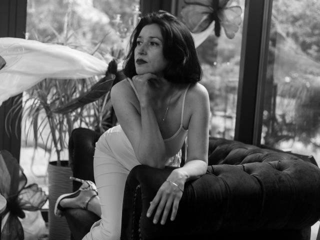 Cameron Enns, con i capelli scuri e il rossetto, è appoggiata a un divano nero imbottito in una foto in bianco e nero, indossa un abito bianco aderente, con la mano vicino alla bocca, e guarda altrove..