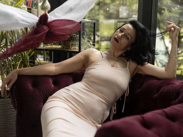CameronEnns is a sexy woman with dark hair, wearing a tight, pale pink dress, reclining on a red velvet couch, her arm raised, eyes closed, and red lips.