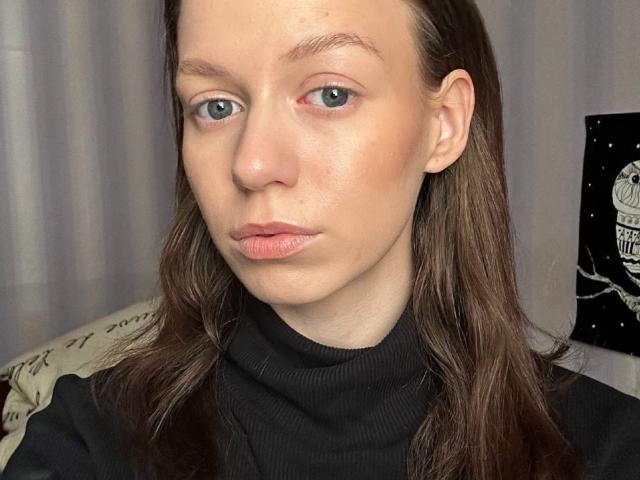 MarnaFantz, met bleke huid, blauwe ogen, bruin haar, gekleed in een zwarte coltrui, heeft lichtroze lippen die iets open zijn, en een kleine rode vlek op haar rechterwang..