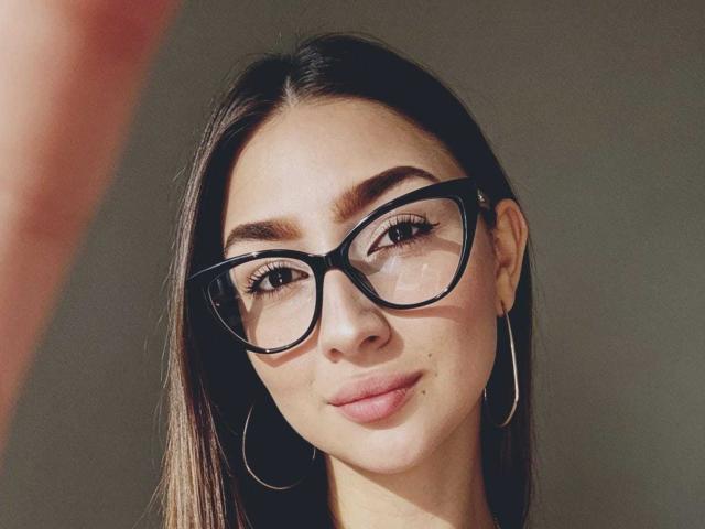 Abby, com óculos pretos grandes, cabelo castanho comprido, brincos de argola e um leve sorriso. Seus seios são pequenos e sua pele é clara. Ela está com uma aparência adorável e sexy..