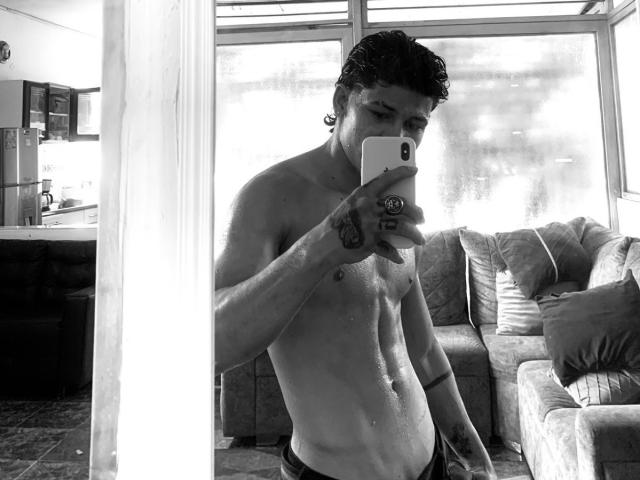 Selfie em preto e branco de TemptKing, sem camisa, com um corpo magro e musculoso, pequenos mamilos e tatuagens, segurando um celular, em frente a um espelho, com um sofá ao fundo..