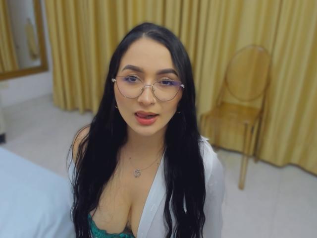 PamelaLiss, con lunghi capelli neri, occhiali e rossetto rosso, indossa un reggiseno verde e un abito bianco, mostrando il seno, seduta in una stanza con tende gialle e una sedia..