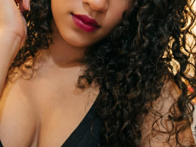 Sadieth tiene el pelo rizado y oscuro, lápiz labial rojo intenso y un top negro sin mangas que deja ver sus pechos. Su mano está cerca de su cabeza. Tono de piel cálido. Ligeramente borroso..