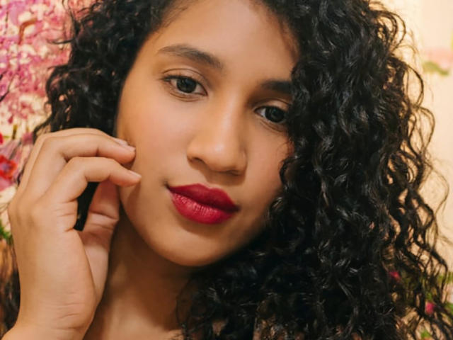 O rosto de Sadieth, com cabelos pretos encaracolados, pele marrom média, batom vermelho, lábios levemente entreabertos, mão tocando a bochecha, expressão suave, luz suave, maquiagem discreta, feminina, atraente..