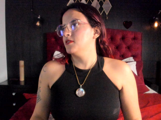 LaiaJonnes met rood haar, een bril, een zwart topje met halter, een gouden ketting en een zilveren hanger, zittend op een rode, gestoffeerde bed, met tatoeages zichtbaar op haar rechterarm..