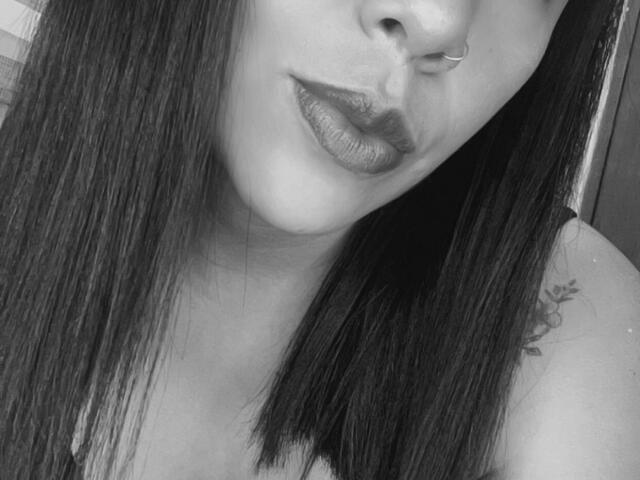 Das Gesicht von Emily Durand auf einem Schwarz-Weiß-Foto, das ihren Nasenring, den glänzenden, dunklen Lippenstift, das glatte, schwarze Haar und ein kleines Tattoo an ihrer rechten Schulter zeigt. Ihre Brüste sind leicht sichtbar..