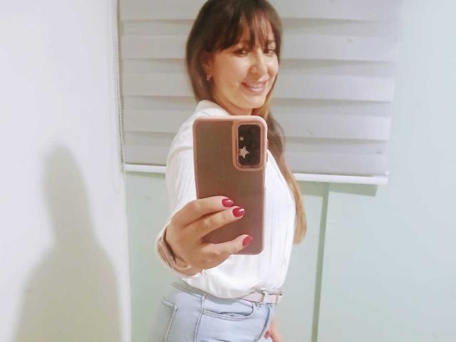 Roxanne Blair, con cabello castaño largo, una blusa blanca y jeans azul claro, sonriendo a sí misma en un selfie frente a un espejo, sosteniendo un teléfono rosa, con uñas rojas, de pie en una habitación..