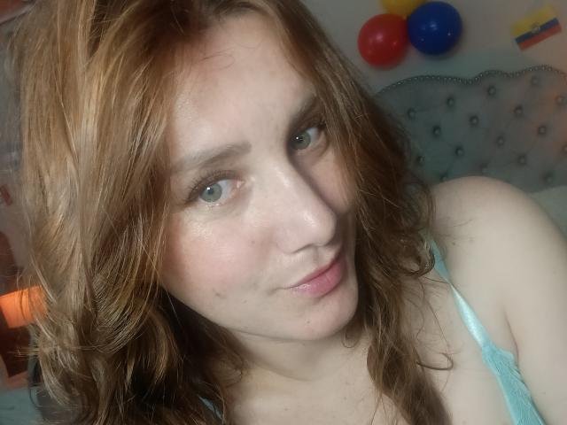 KatyaRogersX with wavy red hair, pale skin, and green eyes, wearing a light blue lace top, smiling softly at the camera. Her tits are partially visible.