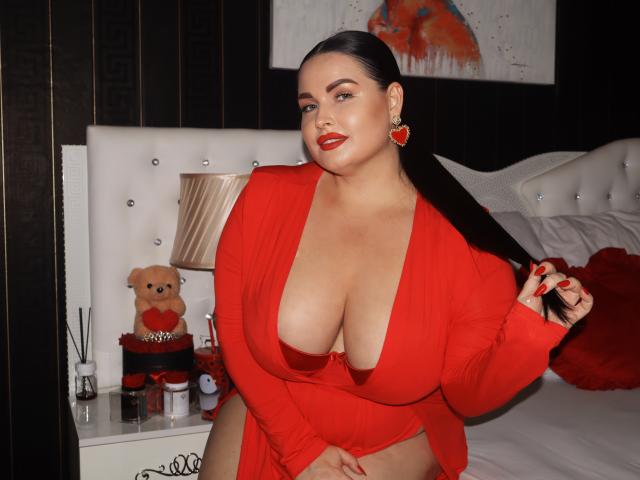 GeorgiaLerox, com seios grandes, vestindo um vestido vermelho, segurando seu rabo de cavalo preto, batom vermelho, brincos vermelhos, sentada em uma cama, um ursinho de pelúcia em cima da mesinha de cabeceira, uma luminária e maquiagem..