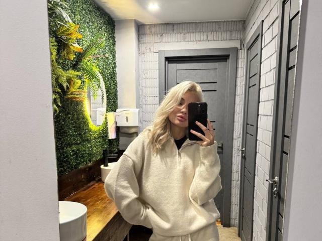 Pearlene Scalley, avec ses cheveux blonds, portant un sweat à capuche blanc duveteux, prend un selfie devant un miroir dans une salle de bain, sa main dans sa poche, tenant un téléphone noir..