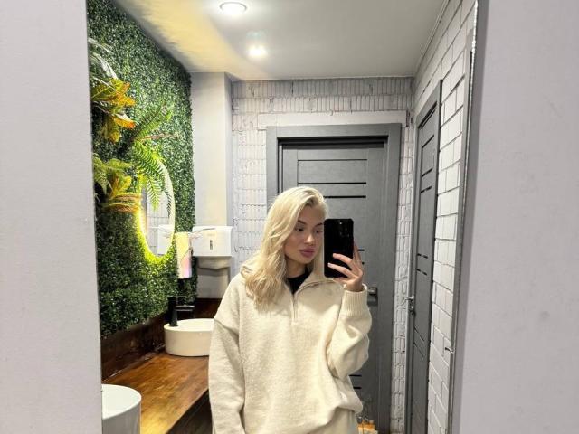 Pearlene Scalley, con i capelli biondi, indossa una felpa bianca e soffice e sta scattando un selfie allo specchio in un bagno con pareti verdi, un mobiletto in legno e una porta grigia. Il seno non è visibile..
